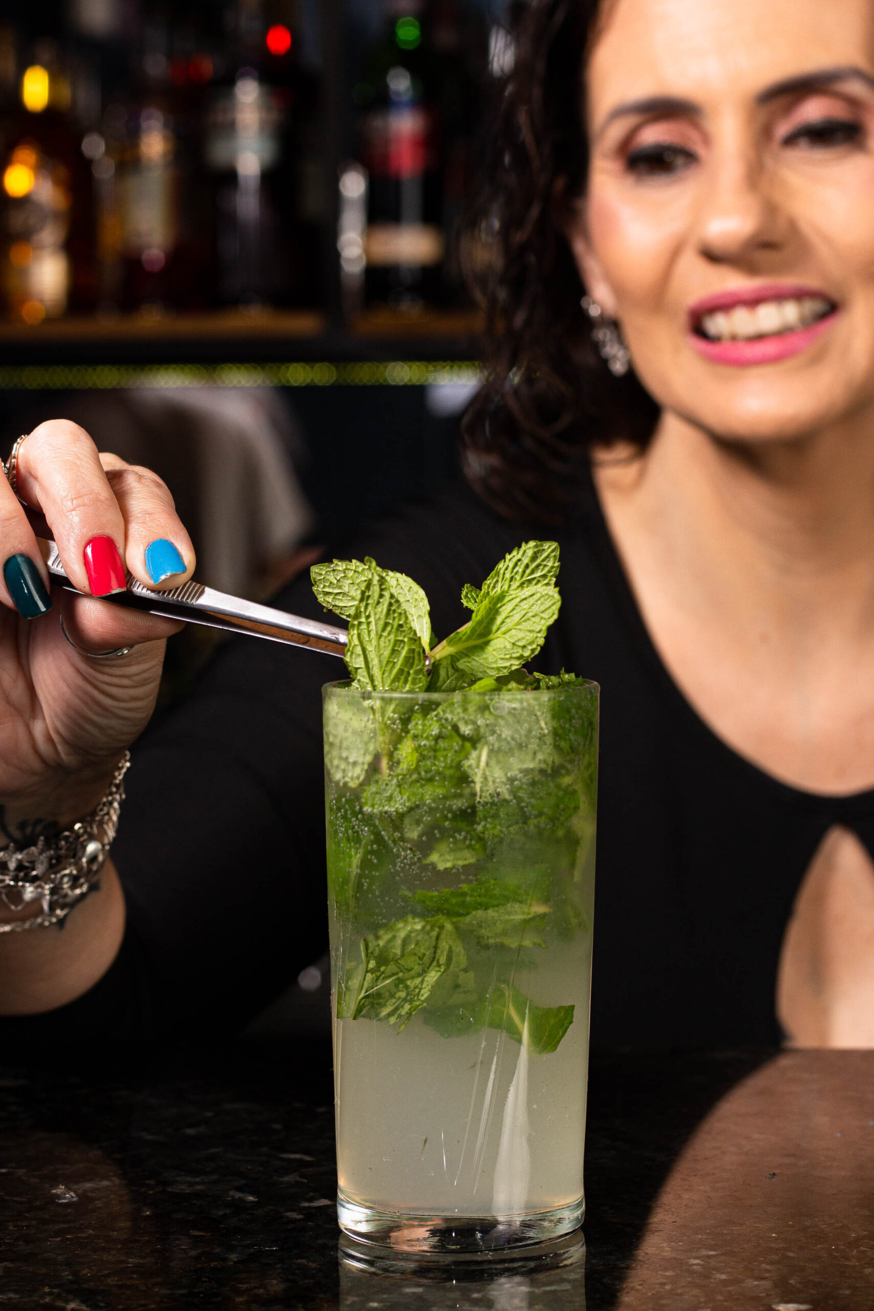 mulher colocando hortelã com uma pinça em um copo com drink.