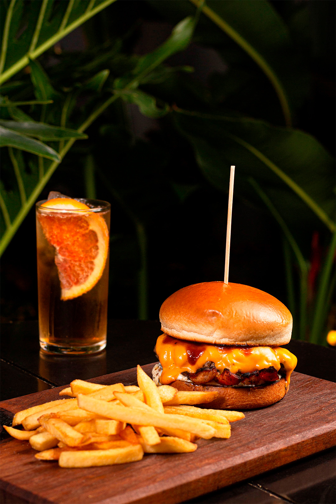 Tábua com batata frita e hamburguer ao fundo copo com drink.