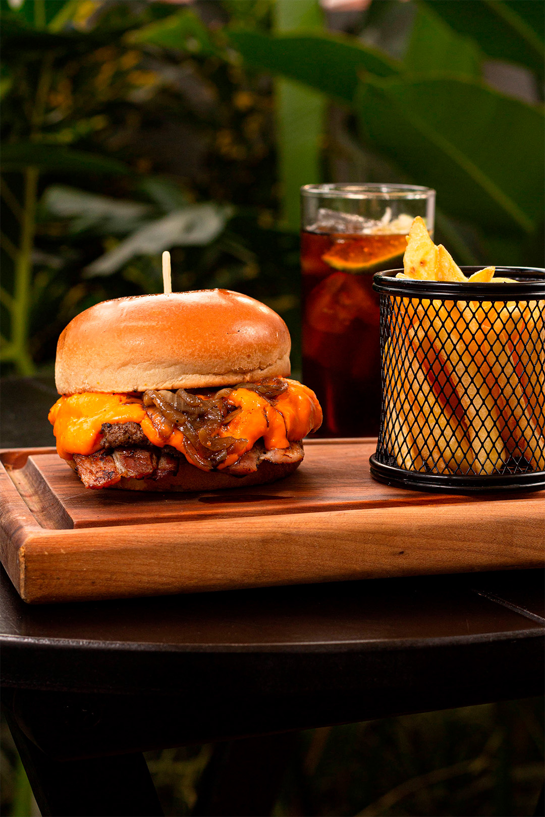 Tábua com hamburguer e batata frita ao fundo copo com refrigerante.