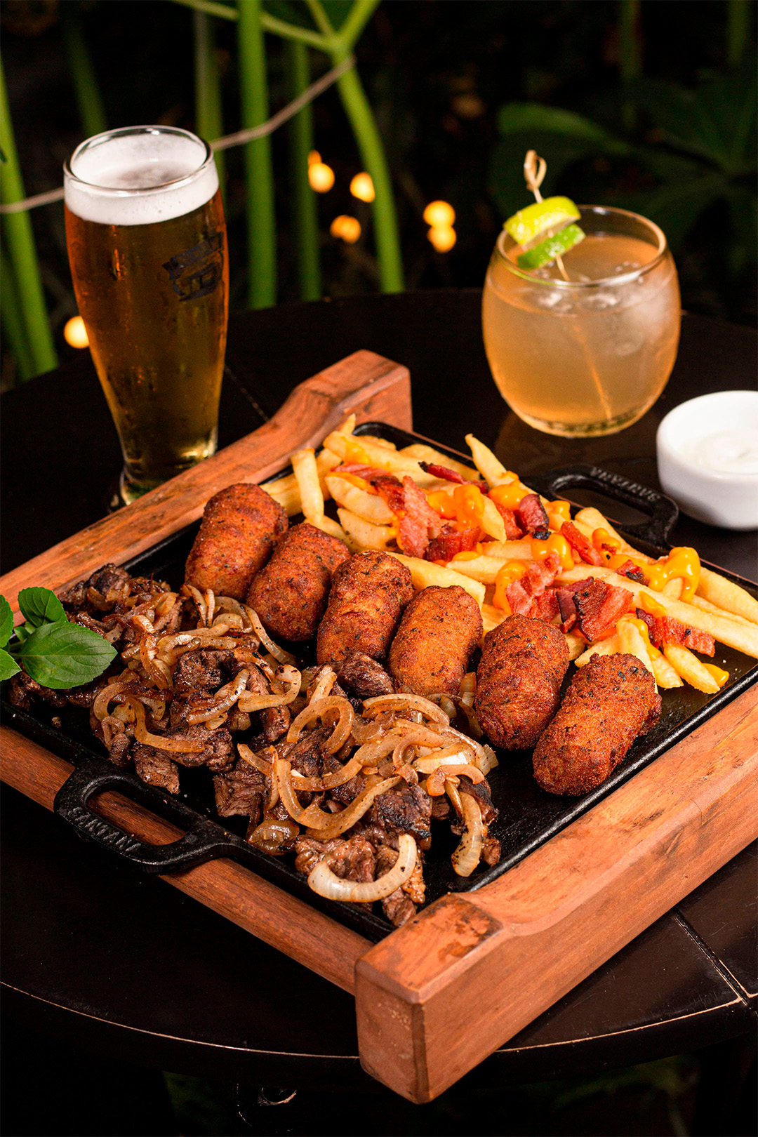 Tábua com porção diversas ao fundo um copo de cerveja e copo com drink.