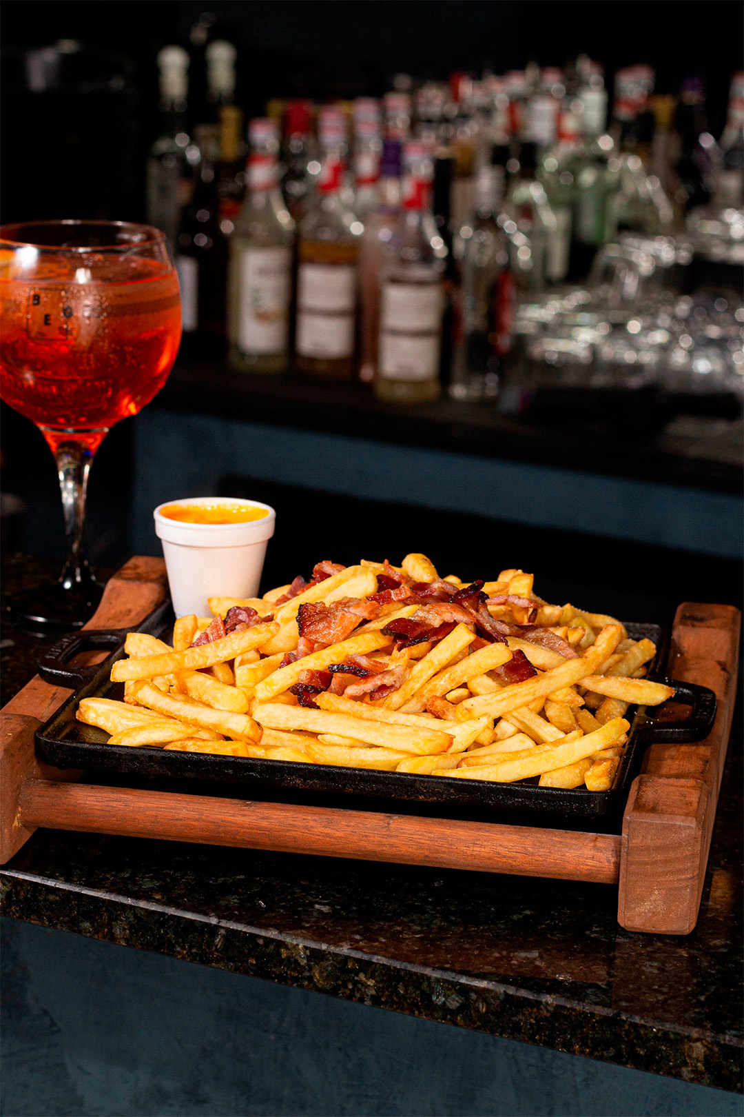 Tábua com porção de batata frita com queijo e bacon ao seu lado um copo com drink.