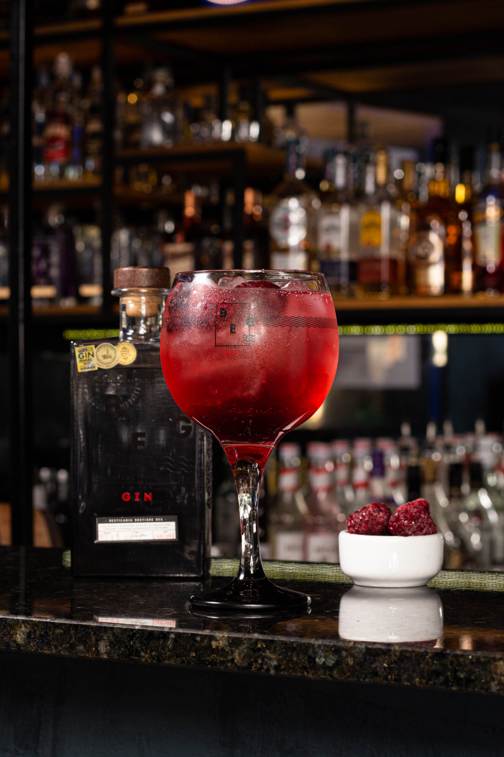 copo de drink ao seu lado uma garrafa de gin e no outro um pote com fruta vermelha