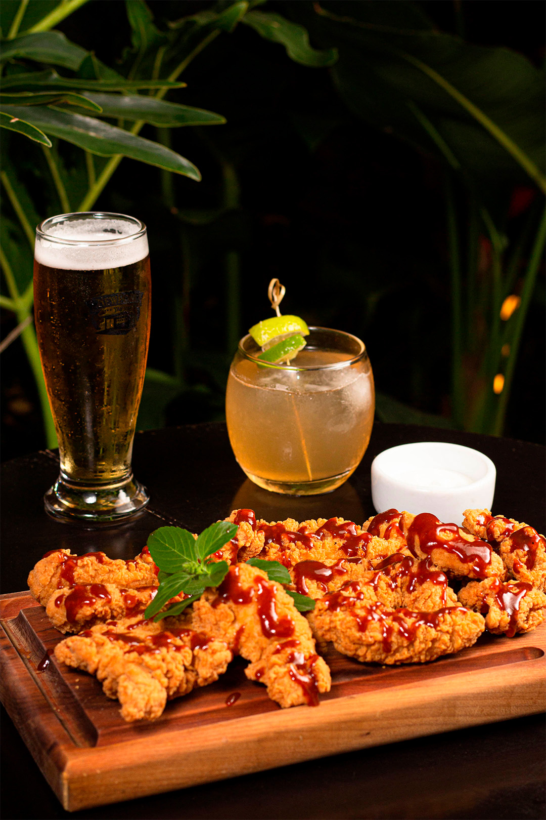 Tábua com stick de frango ao fundo copo com cerveja e como com drink