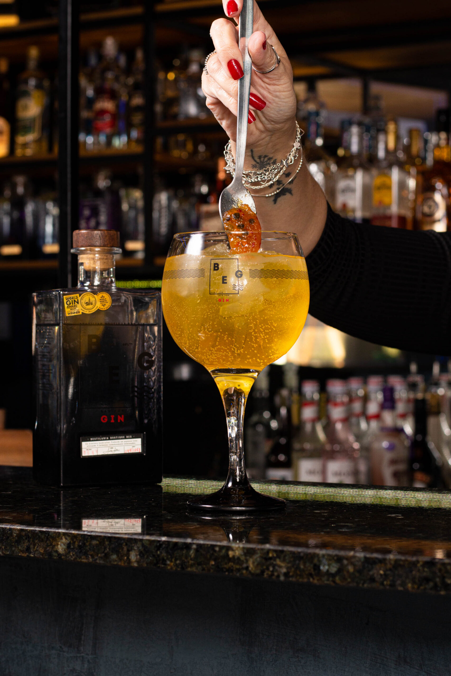 Mulher adicionando maracujá com uma colher ao um copo de drink ao seu lado uma garrafa de gin